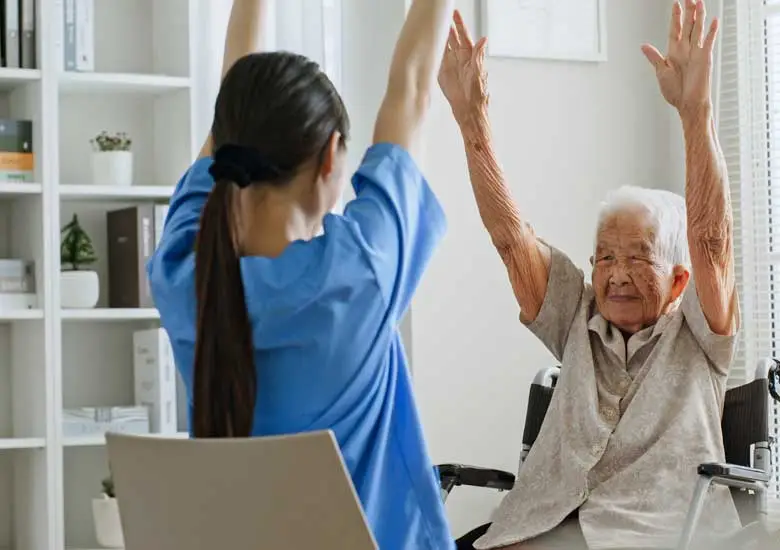 A caregiver is assisting a senior Asian woman with physical therapy exercises for her arms at home